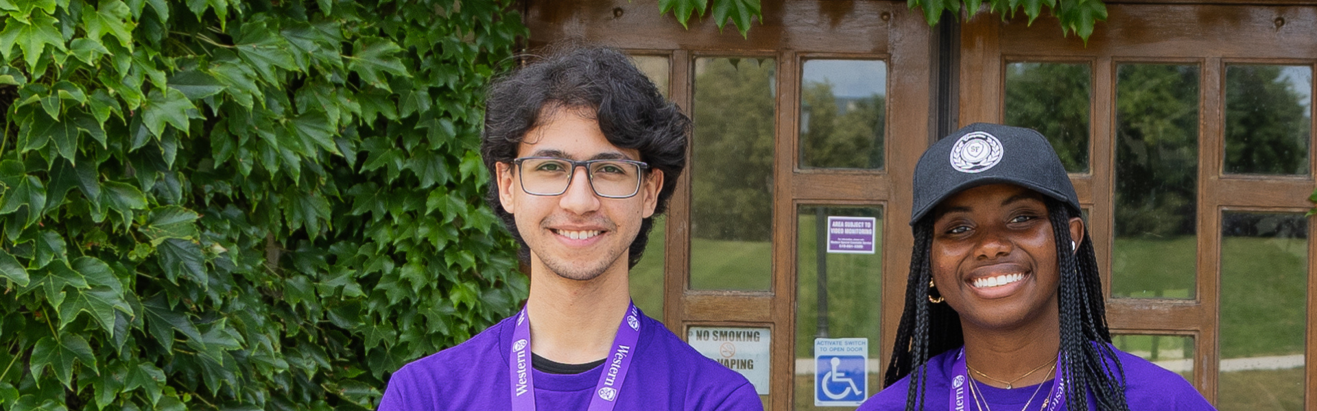 Two student leaders dressed in purple stand in front of a vine-covered building.