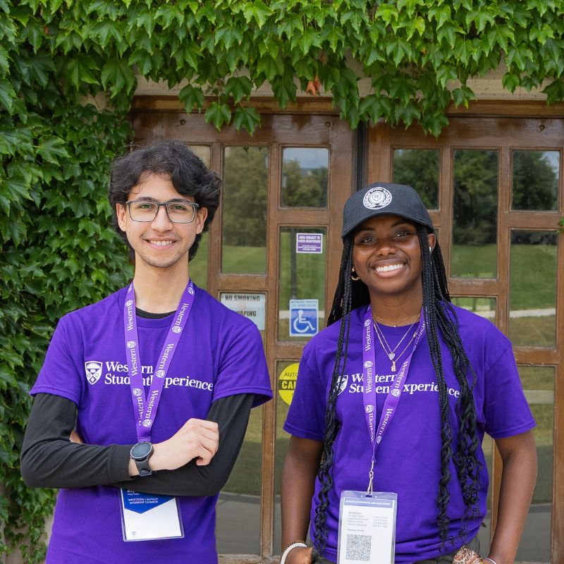 Two student leaders dressed in purple stand in front of a vine-covered building.