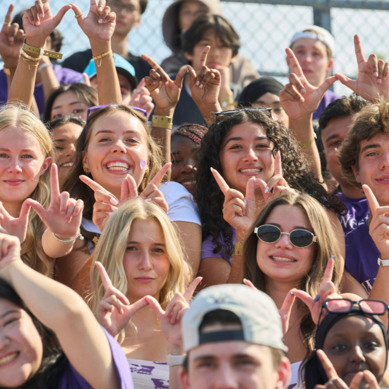 Many students in a crowd dressed in purple, holding up a w-sign with their hands.