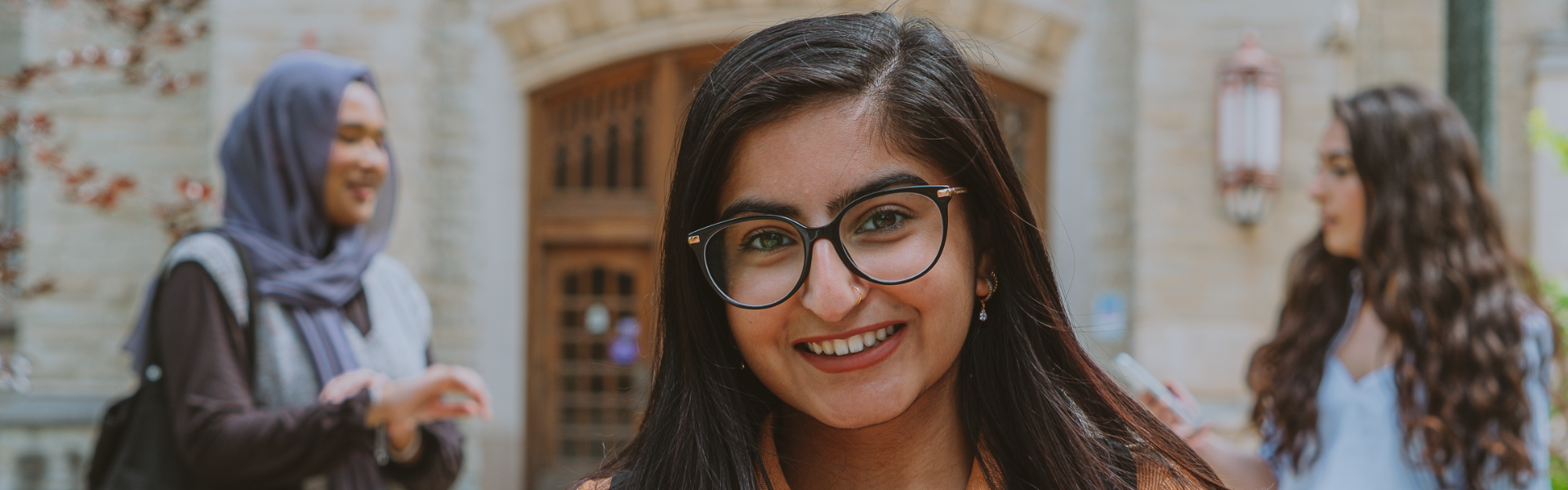 A young woman wearing glasses stands in front of a stone building with two women behind her.