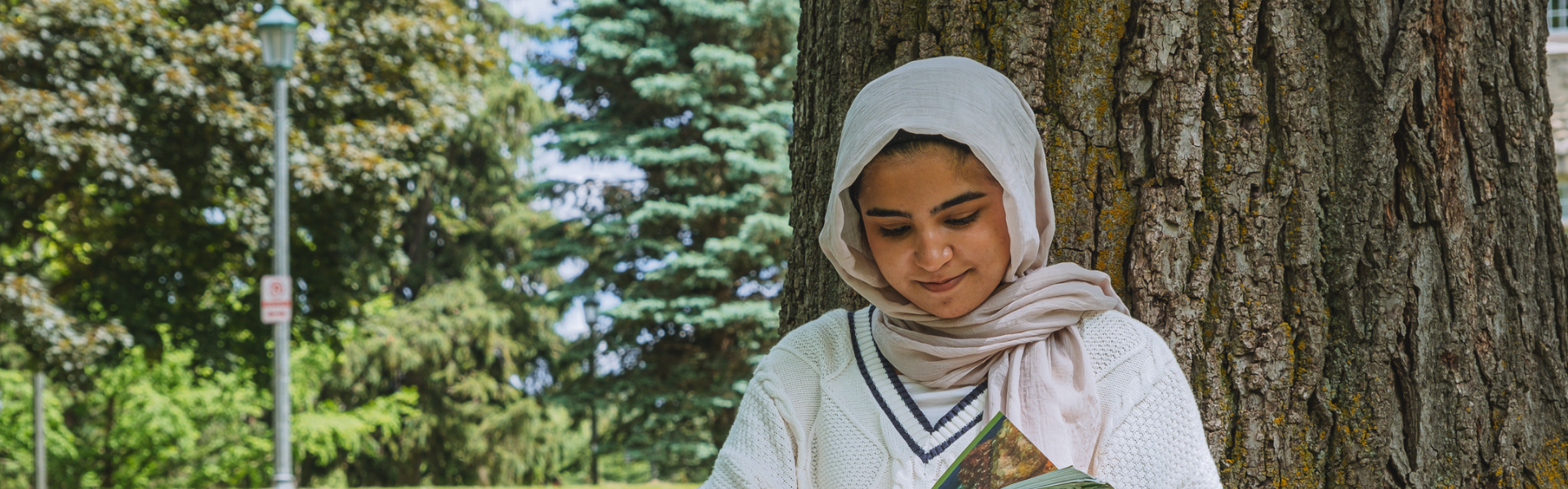 A young woman wearing a hijab studies under a tree.