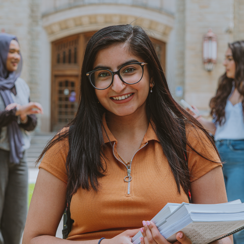A young woman wearing glasses stands in front of a stone building with two women behind her.